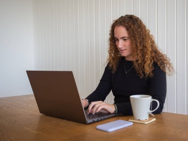 Kızıl saçlı bir kadının elinde bir fincan çayla dizüstü bilgisayar kullanmasının yan görüntüsü.