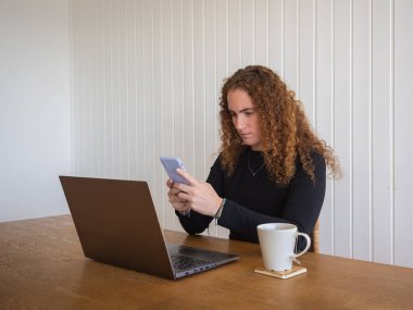 Evde çalışan ve cep telefonunu bir fincan çayla kullanan genç bir kızıl kadının yan görüntüsü.