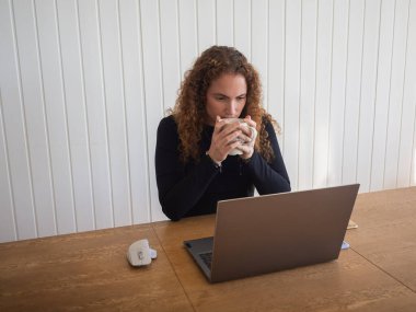 Kızıl saçlı genç kadın bilgisayardan bir şeyler izlerken bir fincan çayla ısınıyor.