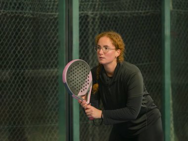 Odaklanmış bir kadın açık hava padel tenis kortunda hizmet etmeye hazırlanıyor, spor kıyafetleri ve ekipmanlar sergileniyor.