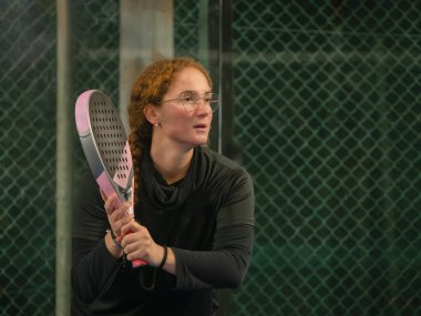 Gözlüklü bir kadın, elinde raketini tutarak Padel tenisine yoğun bir şekilde odaklanır.