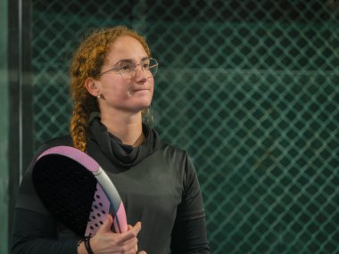 Düşünceli bir kadın padel raketini tutar, açık hava kortuna bakar.