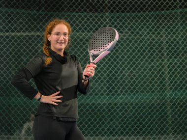 Gülümseyen kadın, açık hava sahasında pembe bir padel raketle rahat bir şekilde poz veriyor.