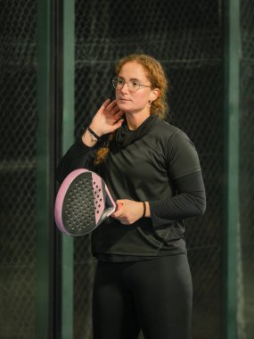 Düşünceli bir kadın padel raketiyle ayakta duruyor, bir sonraki oyununu açık hava maçı sırasında düşünüyor.