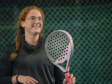Spor kıyafetleri içinde gülümseyen bir kadın, açık bir sahada padel raketi tutuyor, neşe ve neşe saçıyor.