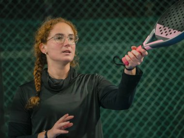 Spor kıyafetleri içindeki bir kadın, açık hava maçında yol tarifi yaparken padel raketiyle güven içinde el kol hareketi yapar.