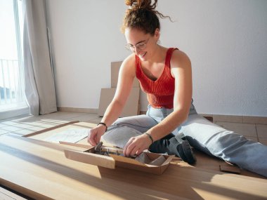 Aletler ve parçalarla çevrili, yere ahşap mobilyalar monte eden neşeli genç kadın.