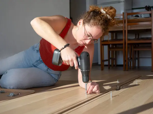 Kablosuz elektrikli matkap kullanarak yere ahşap bir mobilya monte etmeye odaklanmış bir kadın.