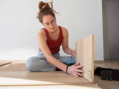 Odaklanmış genç bir kadın yerde otururken ahşap mobilya parçalarını birleştirirken görülüyor.