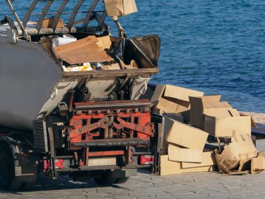 Çöp dolu bir çöp kamyonu, deniz kenarındaki bir gezinti alanında dağınık karton kutuların yanında durup atık imhası çabalarını vurguluyor.