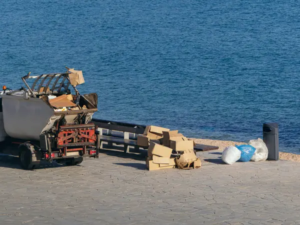 A waste collection truck parked on a seaside promenade, surrounded by piles of cardboard, bags, and other trash, highlighting environmental concerns