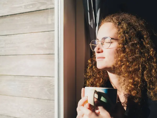 Huzurlu, kıvırcık saçlı ve gözlüklü genç bir kadının yakın çekimi. Pencere kenarında güneşlenirken ılık bir fincan kahvenin tadını çıkarıyor.