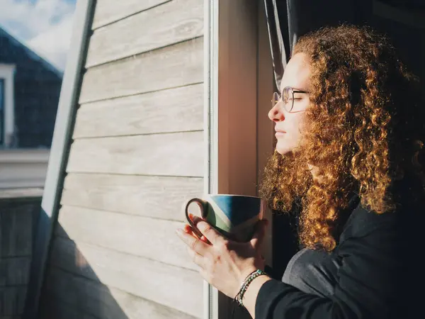 Kıvırcık saçlı bir kadın elinde bir kupa kahveyle pencereden dışarı bakıyor. Rahatlama ve tefekkür kavramı, kadının kahvenin tadını çıkarırken dışarıdaki manzaranın tadını çıkarması gibi.