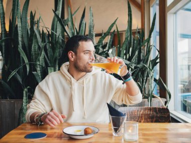 Rahat bir adam, yemyeşil bitkilerle bezenmiş rahat bir kafe ortamında, ahşap bir masada oturan bir bardak birayı yudumluyor.
