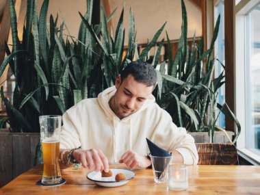 Rahat poz veren genç bir adam, çerezlerinin ve bir bardak birasının keyfini çıkarırken, kafeteryada rahat bir şekilde oturuyordu. Etrafı canlı bitkilerle çevriliydi.