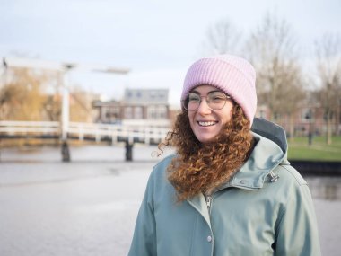 Kıvırcık saçlı, gülümseyen genç bir kadın nehir kenarında güneşli bir kış gününün tadını çıkarıyor. Pembe bere ve deniz mavisi ceket giymiş.