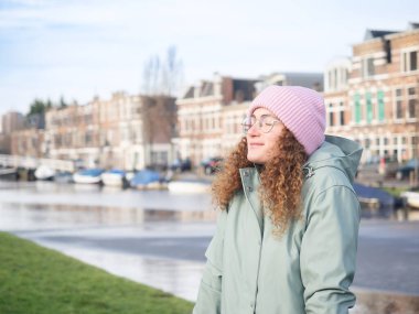 Kıvırcık saçlı, dingin bir genç kadın nehir kenarında bir anlık huzurun tadını çıkarıyor, temiz havayı soluyor, sıcak pembe bir bere ve deniz mavisi ceket giyiyor.