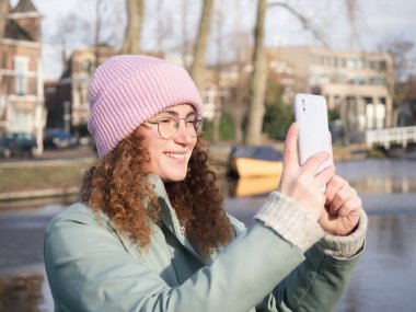 Kıvırcık saçlı neşeli genç bir kadın nehir kenarında pembe bir bere ve açık deniz mavisi ceket giyip Amsterdam 'ın güneşli atmosferinin tadını çıkararak bir fotoğraf çekiyor.