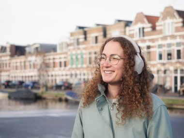Kıvırcık saçlı, gülümseyen, kulaklık takan, tarihi binalar ve arka planda bir kanal olan bir kadın.