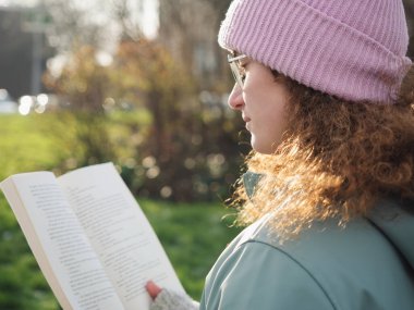 Dalgın, kıvırcık saçlı, gözlüklü ve pembe bereli bir kadının yan görünüşü. Açık havada hafif güneş ışığıyla aydınlatılan bir kitap okumaktan çok etkilenmiş.