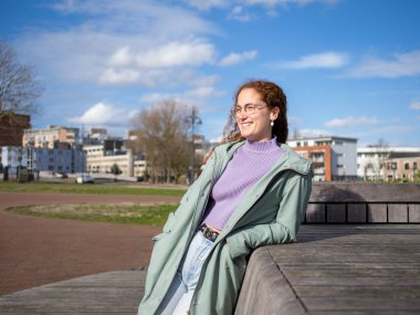 Kıvırcık saçlı, parlak bir gülüşü olan, gözlüklü bir kadın güneşli bir günde Amsterdam 'da bir şehir parkında rahat ve mutlu bir şekilde oturuyor.