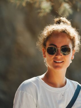 Sıcak bir şekilde gülümseyen genç bir kadının portresi. Spor gözlüğü ve günlük giysiler. Yumuşak güneş ışığıyla işaretlenmiş.