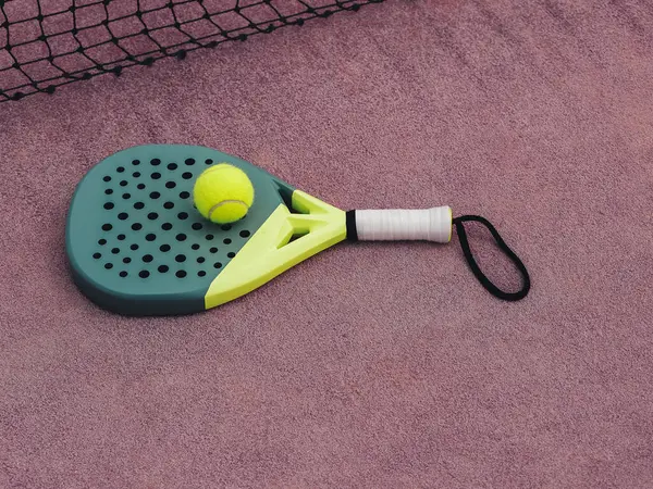 A paddle tennis racket with a neon yellow handle and a matching ball resting on a red clay court