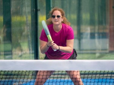 Pembe bluzlu kadın, güneşli bir günde tenis maçında hazır bir pozisyonda çömelmiş topu bekliyor.