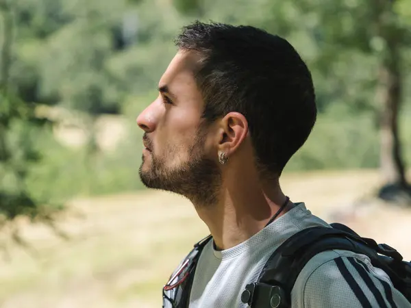 Side profile of a focused male hiker with a backpack, contemplating the scenic view in a forested area