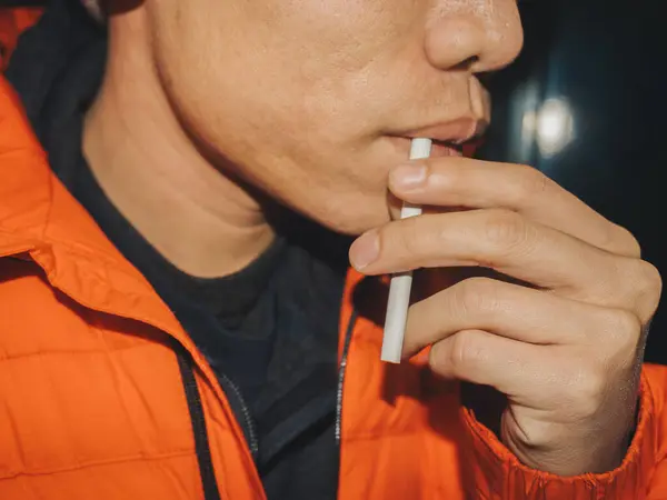 Sigara içen, turuncu ceketli, elleri ve ağzına odaklanmış, elinde sigara tutan Asyalı genç bir adamın detaylı görüntüleri.