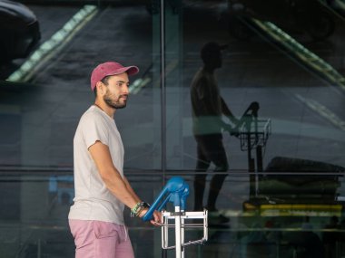 Gündelik giysiler içinde ve kırmızı şapkalı genç bir adam yansıtıcı camlı pencereli havaalanı terminalinin dışına bir bagaj vagonunu itiyor. Seyahat ve ulaşım teması