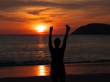 Kutlama için kollarını kaldıran bir insan okyanusun üzerinde canlı bir gün batımını seyrederek akşam gökyüzüne karşı çarpıcı bir siluet yaratıyor.