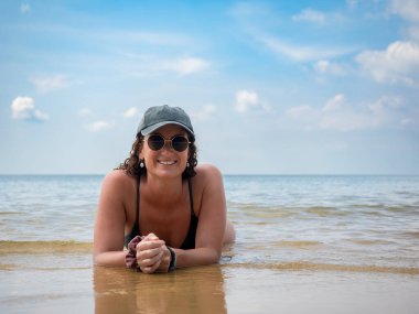 Şapkalı ve bikinili bir kadın, geniş bir gülümsemeyle güneşli bir günde sığ okyanus suyunda uzanıyor. Sakin deniz ve mavi gökyüzü onun mutluluğu için mükemmel bir zemin oluşturur.