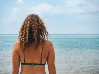 Kıvırcık saçlı bir kadın sakin okyanusa bakıyor, bikini giyiyor. Huzurlu sahil ve berrak gökyüzü huzurlu ve düşünceli bir an sunar.