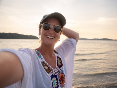 Güneş gözlüklü neşeli bir kadın ve renkli bir üst günbatımında tropik bir plajda selfie çeken sakin okyanus dalgaları ve arka planda bir ada olan bir kadın.