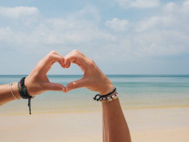 Bilezikli ellerin yakın çekimi berrak mavi gökyüzü ve sakin okyanus sularıyla sakin bir tropik plajın arka planına karşı bir kalp şekli oluşturur.