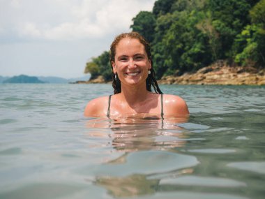 Islak saçlı bir kadın, yemyeşil bir sahil şeridinin yakınındaki berrak okyanus sularında yüzerken gülümsüyor. Sakin ortam ve sakin sular rahatlatıcı bir atmosfer yaratır.