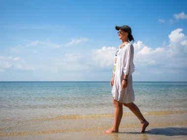 Beyaz örtülü ve şapkalı bir kadın huzurlu bir plajda yalınayak yürüyor, güneşli bir günde hafif dalgaların ve açık mavi gökyüzünün tadını çıkarıyor.