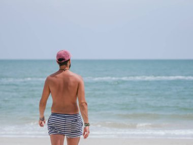 Çizgili şort ve pembe şapkalı bir adamın kumlu bir sahilde durduğu, berrak bir gökyüzünün altındaki sakin okyanusu izlediği arka plan.