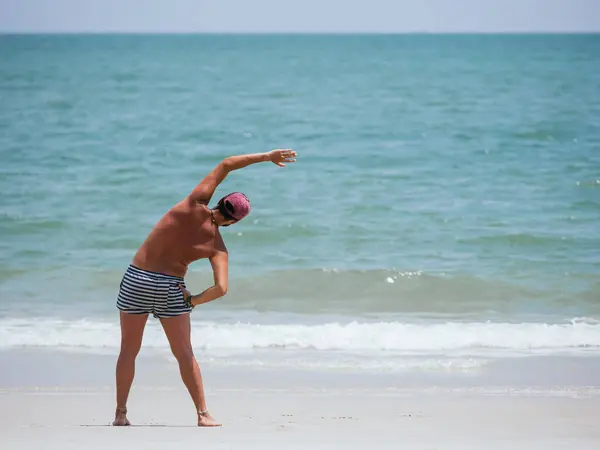 stock image A man wearing striped shorts and a red cap stretches his arms on a sandy beach with the ocean in the background. The sky is clear and the water is calm