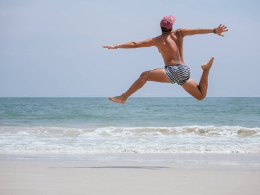 Çizgili şortlu ve pembe şapkalı bir adam kumlu bir sahilde coşkuyla zıplıyor, mavi okyanus ve arka planda hafif dalgalar, neşe ve özgürlüğü ifade ediyor.