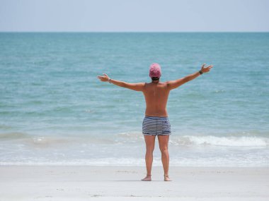 Çizgili şortlu ve şapkalı bir adam kollarını okyanusa doğru açmış güneşli bir plaj gününde özgürlüğünü ve rahatlığını simgeleyen tatlı deniz esintisini kucaklıyor.