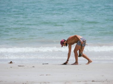 Çizgili şortlu ve şapkalı bir adam sahil boyunca enkaz toplayarak sahili temizliyor, sakin bir kıyı şeridinde çevresel bakım ve idarecilik gösteriyor.