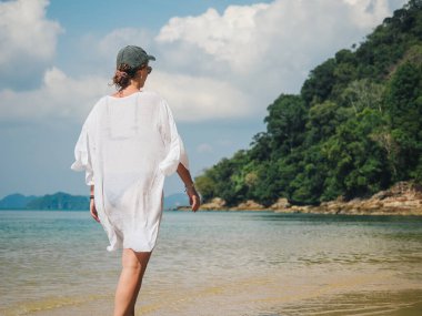 Beyaz örtülü ve şapkalı genç bir kadın yemyeşil yeşillikler ve berrak mavi sularla çevrili sakin tropikal bir sahil boyunca yürüyor.