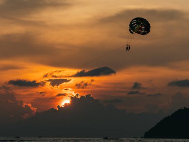 Okyanusun yukarısında deniz paraşütü yapan bir çift dramatik ve canlı bir günbatımı gökyüzüne karşı siluet oluşturarak romantik ve maceralı bir sahne yaratıyor. Alacakaranlık gökyüzünde romantik macera kavramı