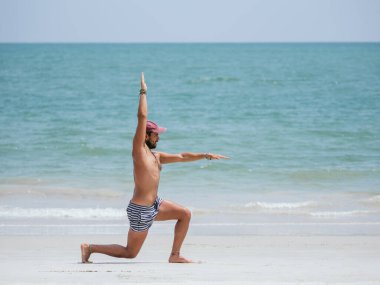 Çizgili şortlu ve şapkalı bir adam kumlu bir plajda savaşçı yoga pozu veriyor. Arkaplanı olarak okyanusla uyum ve gücü somutlaştırıyor.