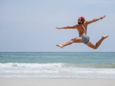 Çizgili yüzme şortu ve şapkası olan bir adam kollarını ve bacaklarını kumlu bir plaja uzatmış güneşli bir günde neşe ve özgürlüğü ifade ediyor.