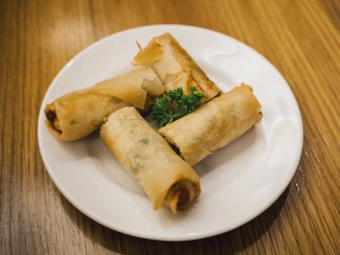 Beyaz bir tabakta servis edilen, taze maydanozla süslenmiş, ahşap bir masaya yerleştirilmiş dört çıtır böreğin yakın çekimi. İştah açıcı Asya atıştırması