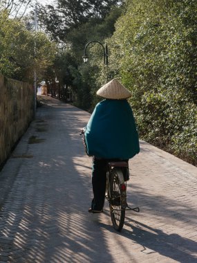 Geleneksel bir şapka takan biri, yemyeşil ve ahşap çitlerle çevrili, güneşli bir yolda bisiklet sürüyor ve huzurlu bir kültürel yolculuğun altını çiziyor.