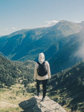 Kapüşonlu ve sırt çantalı tek başına bir yürüyüşçü bir kayanın üzerinde duruyor, geniş bir dağ vadisine tepeden bakıyor Pireneler 'de orman yamaçları ve açık gökyüzü var - doğada bir başarı duygusu yaşıyor.